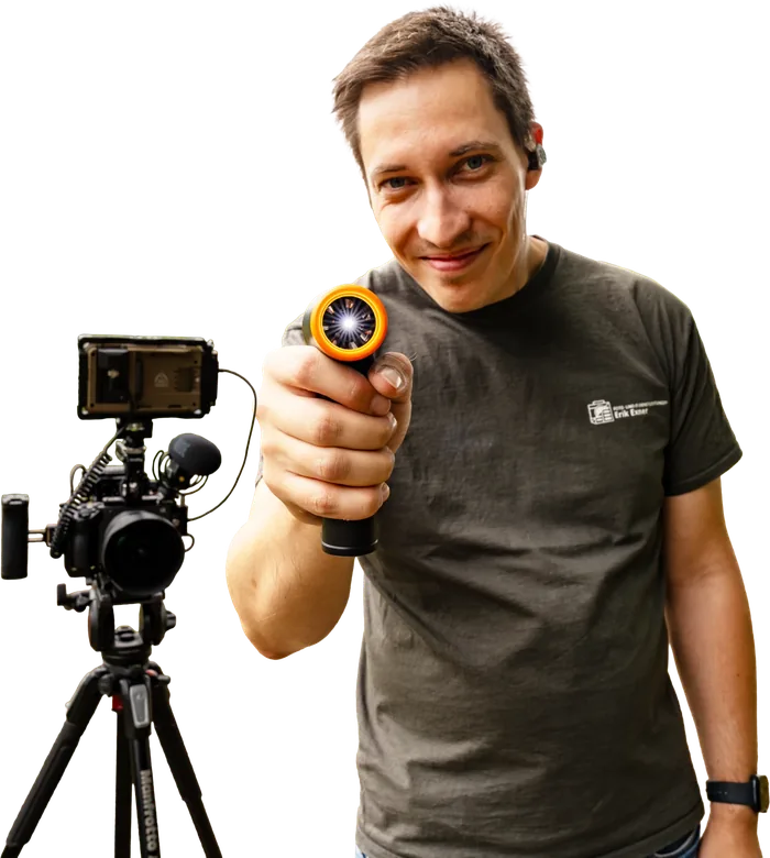 Erik Exner neben seiner Videokamera auf dem Stativ, leuchtet grinsend mit einer Lampe in Richtung Betrachter